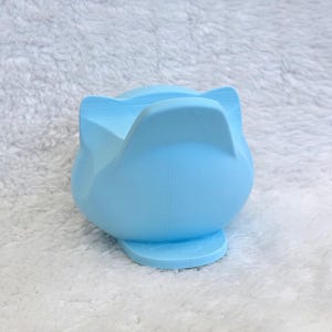 May include: A light blue silicone phone stand shaped like a cat head with pointed ears. The object features a smooth, matte finish and a wide base for stability, designed to hold a mobile device in an upright position for viewing.