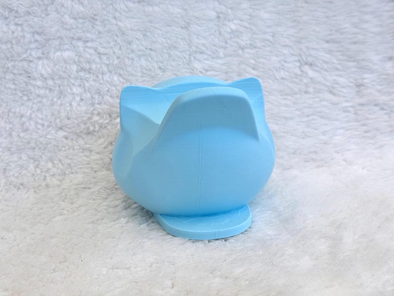 May include: A light blue silicone phone stand shaped like a cat head with pointed ears. The object features a smooth, matte finish and a wide base for stability, designed to hold a mobile device in an upright position for viewing.