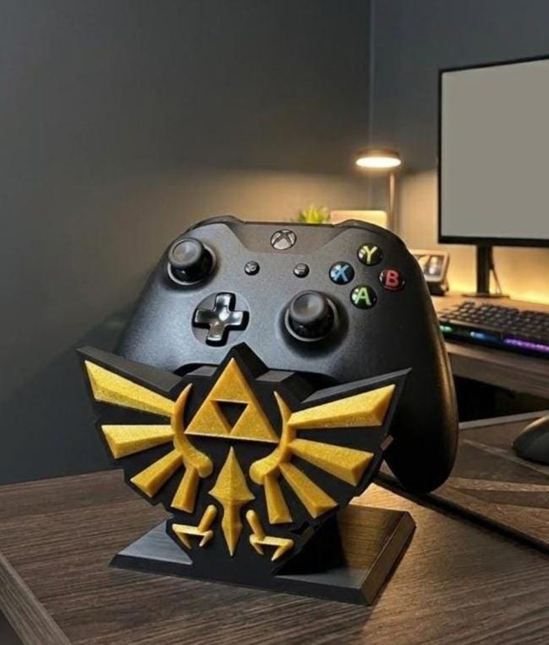 May include: A black Xbox controller rests on a custom-made stand featuring the gold and black emblem of the Legend of Zelda. The stand is on a wooden desk, with a computer monitor and keyboard in the background.