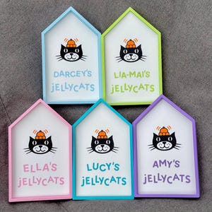 May include: Five house-shaped signs with colorful frames. Each sign features a black cat illustration wearing a pumpkin hat and the text "DARCEY'S JELLYCATS", "LIA-MAI'S JELLYCATS", "ELLA'S JELLYCATS", "LUCY'S JELLYCATS", and "AMY'S JELLYCATS" in different colors.