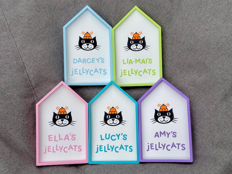 May include: Five house-shaped signs with colorful frames. Each sign features a black cat illustration wearing a pumpkin hat and the text "DARCEY'S JELLYCATS", "LIA-MAI'S JELLYCATS", "ELLA'S JELLYCATS", "LUCY'S JELLYCATS", and "AMY'S JELLYCATS" in different colors.