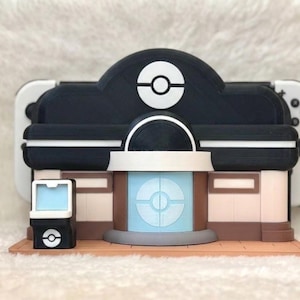 May include: A multi-colored 3D-printed model of a Pok&eacute;mon Center, featuring a black and white color scheme with a light blue central element. The model includes a Pok&eacute; Ball logo and is designed to hold a gaming console.