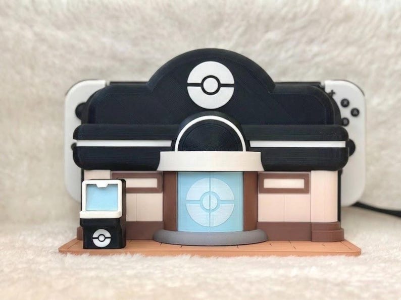 May include: A multi-colored 3D-printed model of a Pok&eacute;mon Center, featuring a black and white color scheme with a light blue central element. The model includes a Pok&eacute; Ball logo and is designed to hold a gaming console.