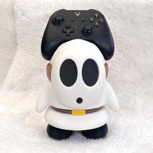 May include: A white Shy Guy character figure serving as a controller stand, featuring a black mask with two large oval eye holes and a small circular mouth. The figure wears a brown belt with a yellow buckle and black shoes, supporting a black game controller.