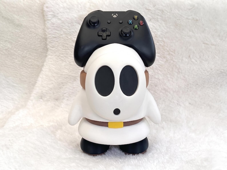 May include: A white Shy Guy character figure serving as a controller stand, featuring a black mask with two large oval eye holes and a small circular mouth. The figure wears a brown belt with a yellow buckle and black shoes, supporting a black game controller.