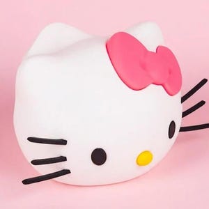 May include: A white, three-dimensional Hello Kitty figure with a pink bow, black whiskers, and black and yellow facial features. The figure is set against a pink background.