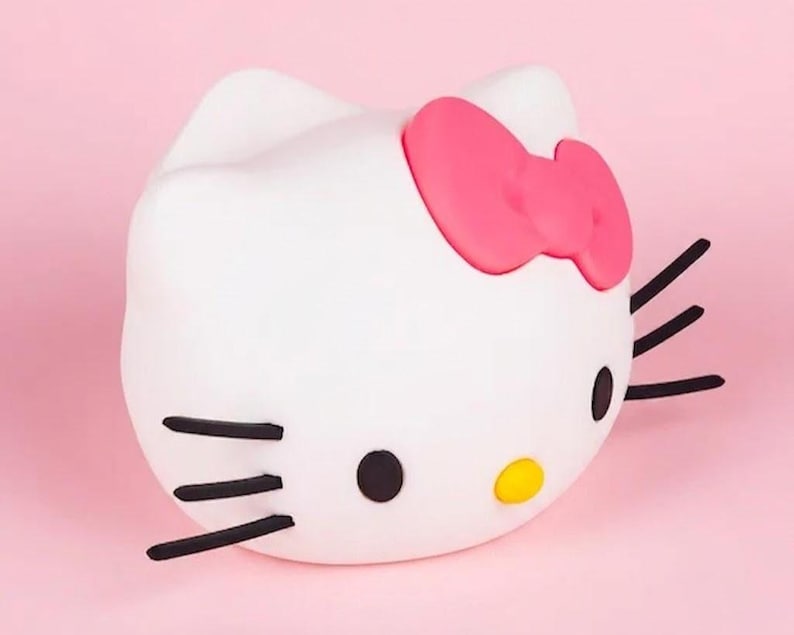 May include: A white, three-dimensional Hello Kitty figure with a pink bow, black whiskers, and black and yellow facial features. The figure is set against a pink background.