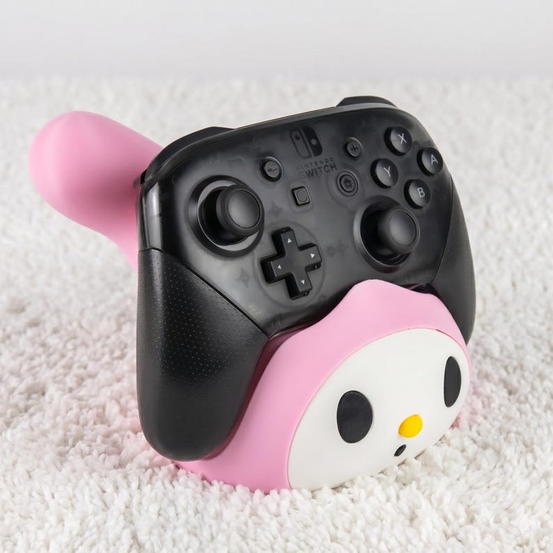 May include: A black Nintendo Switch Pro controller resting in a pink and white character-themed stand. The controller features a translucent black shell, two black analog sticks, a directional pad, and standard action buttons labeled A, B, X, and Y.