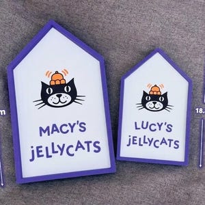May include: Two house-shaped signs with a white background and purple trim. Each sign features a black cat illustration with an orange hat and the text "MACY'S JELLYCATS" and "LUCY'S JELLYCATS". The larger sign is 26.2 cm tall.