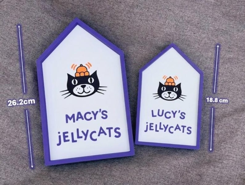 May include: Two house-shaped signs with a white background and purple trim. Each sign features a black cat illustration with an orange hat and the text "MACY'S JELLYCATS" and "LUCY'S JELLYCATS". The larger sign is 26.2 cm tall.