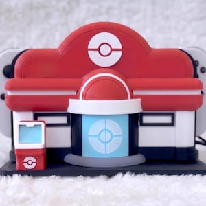 May include: A red, white, and blue plastic Nintendo Switch dock shaped like a Pok&eacute;mon Center building. The structure features a central Pok&eacute; Ball emblem on the roof, a light blue door with a circular design, and a small matching red kiosk on the left.