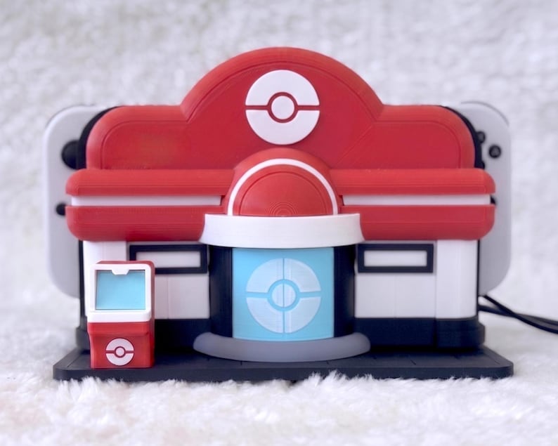 May include: A red, white, and blue plastic Nintendo Switch dock shaped like a Pok&eacute;mon Center building. The structure features a central Pok&eacute; Ball emblem on the roof, a light blue door with a circular design, and a small matching red kiosk on the left.