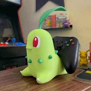 May include: A lime green Chikorita-shaped controller holder with a black controller inserted. The holder has red eyes, green spikes, and a green leaf. A small yellow Pikachu figure is visible in the background.