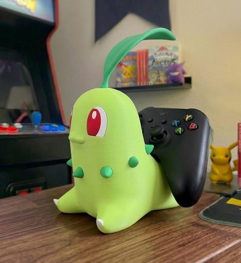 May include: A lime green Chikorita-shaped controller holder with a black controller inserted. The holder has red eyes, green spikes, and a green leaf. A small yellow Pikachu figure is visible in the background.