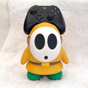 May include: A yellow and green character figurine with a white oval mask featuring two large black eye holes and a small black mouth. A black wireless game controller is positioned on top of the character's head, serving as a functional stand.