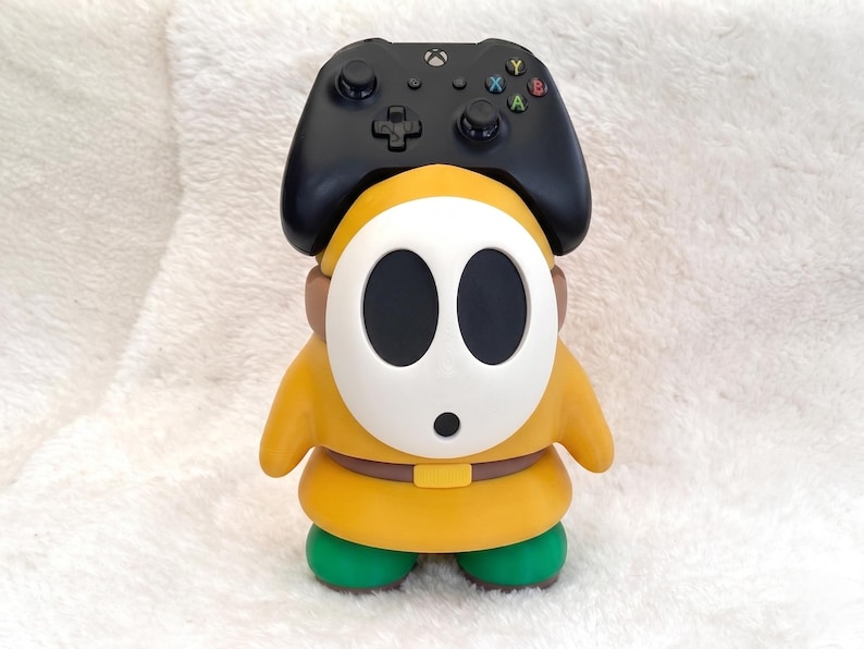 May include: A yellow and green character figurine with a white oval mask featuring two large black eye holes and a small black mouth. A black wireless game controller is positioned on top of the character's head, serving as a functional stand.
