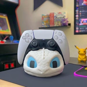 May include: A white and black PlayStation 5 controller sits atop a white dragon-themed controller stand with blue eyes. The stand is on a dark grey surface, with a Pikachu figure and a Game Boy game case in the background.