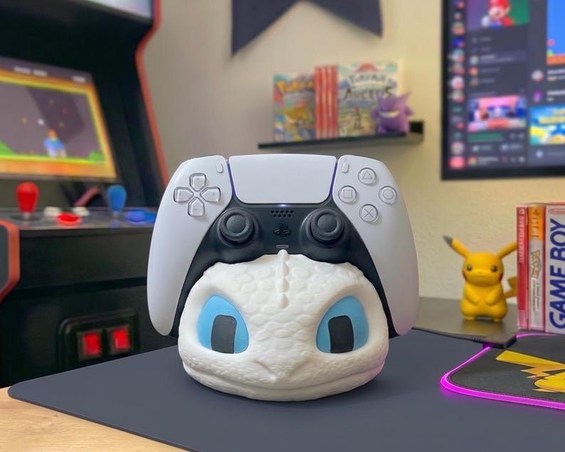 May include: A white and black PlayStation 5 controller sits atop a white dragon-themed controller stand with blue eyes. The stand is on a dark grey surface, with a Pikachu figure and a Game Boy game case in the background.