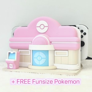 Puede incluir: Modelo rosa y blanco impreso en 3D de un edificio con un diseño circular en el techo y una máquina recreativa rosa más pequeña. Un mando blanco de Nintendo Switch es visible detrás del edificio. El texto "+ FREE Funsize Pokemon" está en la parte inferior.