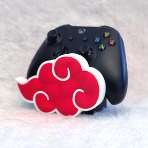 May include: A black video game controller resting behind a three-dimensional red cloud-shaped stand with a thick white border. The controller features standard black joysticks, a directional pad, and four colored action buttons labeled A, B, X, and Y.
