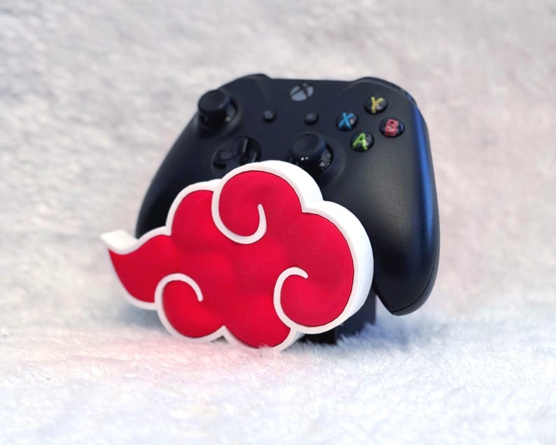 May include: A black video game controller resting behind a three-dimensional red cloud-shaped stand with a thick white border. The controller features standard black joysticks, a directional pad, and four colored action buttons labeled A, B, X, and Y.