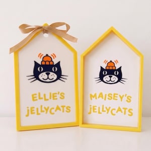 Puede incluir: Dos letreros de madera en forma de casa con marcos amarillos y centros blancos. Cada letrero presenta una ilustraci&oacute;n de un gato negro con un sombrero naranja y el texto "ELLIE'S JELLYCATS" o "MAISEY'S JELLYCATS" en amarillo. El letrero de la izquierda tiene un lazo dorado.