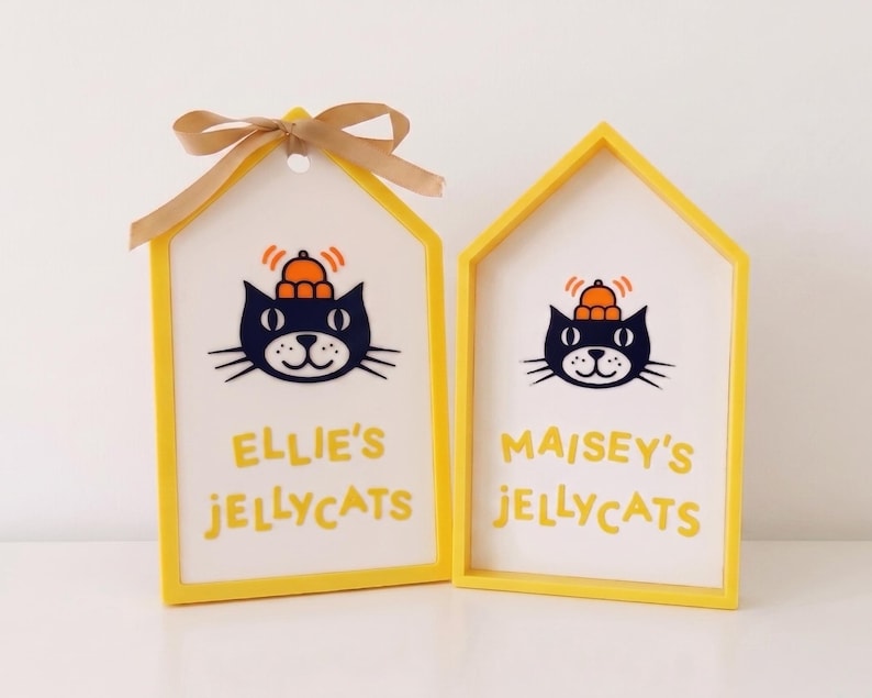 Puede incluir: Dos letreros de madera en forma de casa con marcos amarillos y centros blancos. Cada letrero presenta una ilustraci&oacute;n de un gato negro con un sombrero naranja y el texto "ELLIE'S JELLYCATS" o "MAISEY'S JELLYCATS" en amarillo. El letrero de la izquierda tiene un lazo dorado.
