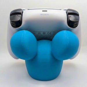 May include: A blue 3D-printed controller stand shaped like a cartoon character, holding a white PlayStation 4 controller. The stand has two large, round ears and a rounded base. The controller is white with black joysticks.