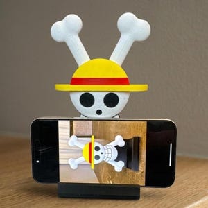 May include: A phone stand featuring a white skull with crossbones and a yellow hat with a red stripe. The stand is holding a smartphone displaying the same skull and crossbones design. The stand is black.