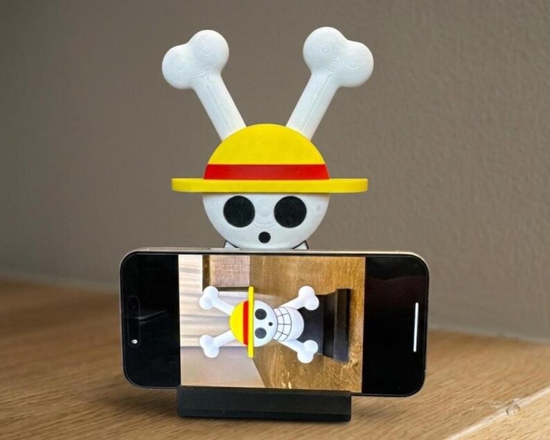 May include: A phone stand featuring a white skull with crossbones and a yellow hat with a red stripe. The stand is holding a smartphone displaying the same skull and crossbones design. The stand is black.