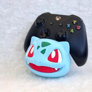 May include: A black Xbox video game controller resting behind a light blue Bulbasaur character stand. The stand features a wide red mouth, white teeth, and dark green spots on the forehead, providing a stable base for the gaming controller.