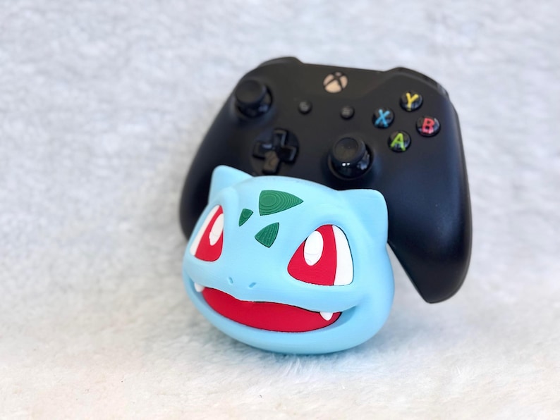 May include: A black Xbox video game controller resting behind a light blue Bulbasaur character stand. The stand features a wide red mouth, white teeth, and dark green spots on the forehead, providing a stable base for the gaming controller.