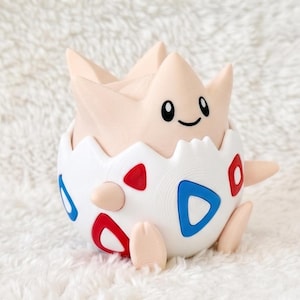 May include: A light beige creature with a rounded head and pointed ears emerging from a white egg-shaped shell. The shell features a pattern of red and blue triangular shapes. The creature has black oval eyes and a small black curved mouth.