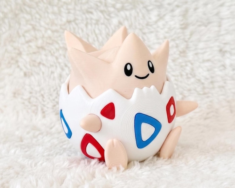 May include: A light beige creature with a rounded head and pointed ears emerging from a white egg-shaped shell. The shell features a pattern of red and blue triangular shapes. The creature has black oval eyes and a small black curved mouth.