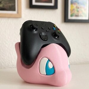 May include: A black Xbox controller rests on a pink 3D-printed Pok&eacute;mon character-themed stand. The stand features a cartoon-style design with blue and white accents. The controller has multiple buttons and joysticks.