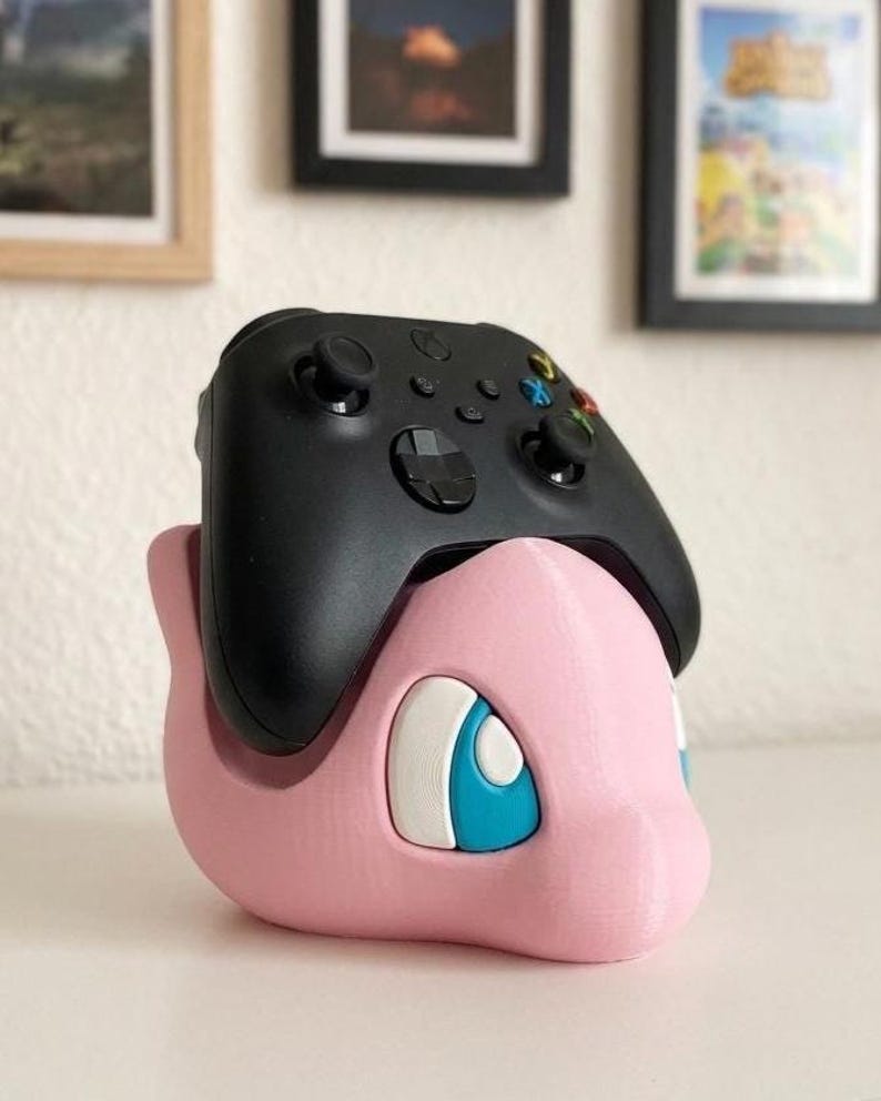 May include: A black Xbox controller rests on a pink 3D-printed Pok&eacute;mon character-themed stand. The stand features a cartoon-style design with blue and white accents. The controller has multiple buttons and joysticks.