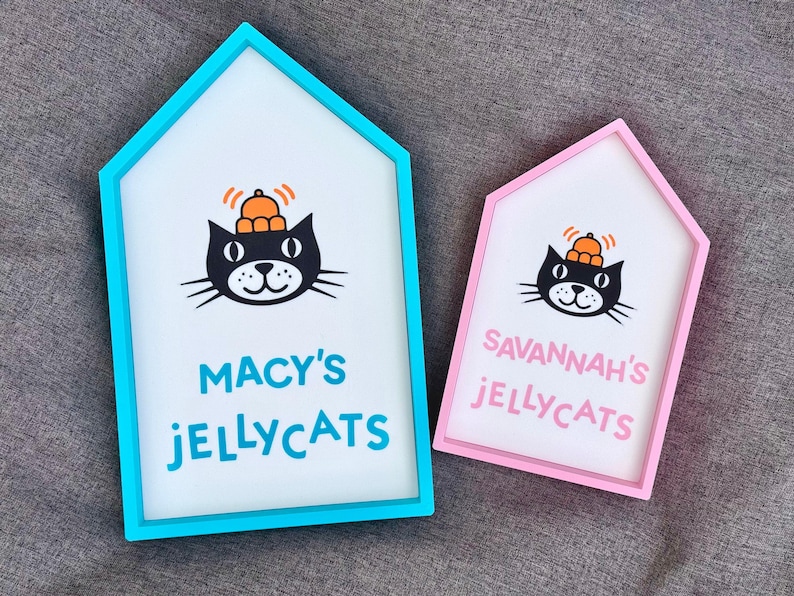 May include: Two house-shaped signs with white backgrounds and colorful frames. Each sign features a black cat illustration wearing an orange hat. One sign reads "MACY'S JELLYCATS" in blue, the other reads "SAVANNAH'S JELLYCATS" in pink.