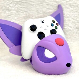 Puede incluir: Un soporte para mando con forma de criatura morada y lila, con grandes orejas puntiagudas y un detalle circular rojo en la frente. Un mando inal&aacute;mbrico Xbox blanco con joysticks negros y botones de colores descansa sobre el soporte.