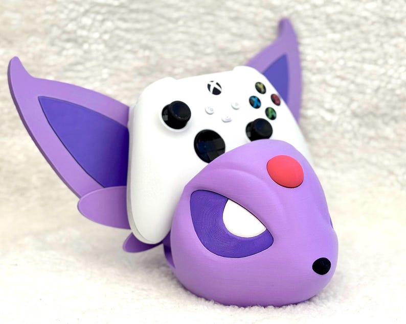 Puede incluir: Un soporte para mando con forma de criatura morada y lila, con grandes orejas puntiagudas y un detalle circular rojo en la frente. Un mando inal&aacute;mbrico Xbox blanco con joysticks negros y botones de colores descansa sobre el soporte.