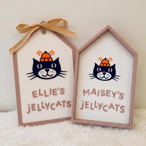 Puede incluir: Dos letreros de madera en forma de casa con marcos rosa claro y centros blancos. Cada uno muestra una ilustraci&oacute;n de un gato negro con un sombrero naranja, con el texto "ELLIE'S JELLYCATS" y "MAISEY'S JELLYCATS" en rosa. Un lazo dorado est&aacute; atado al primer letrero.