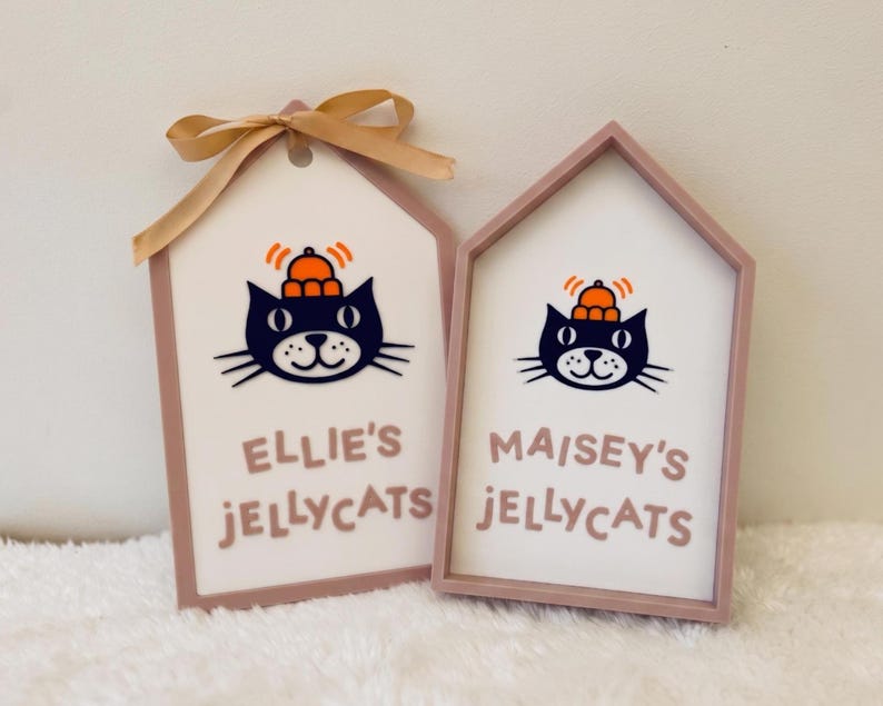 Puede incluir: Dos letreros de madera en forma de casa con marcos rosa claro y centros blancos. Cada uno muestra una ilustraci&oacute;n de un gato negro con un sombrero naranja, con el texto "ELLIE'S JELLYCATS" y "MAISEY'S JELLYCATS" en rosa. Un lazo dorado est&aacute; atado al primer letrero.