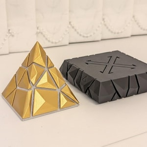 May include: A gold-colored geometric pyramid puzzle and a black, angular, square-shaped device with a cross design. The pyramid is made of triangular sections. The device has a cable attached.