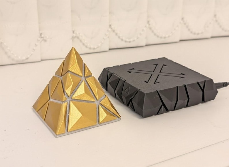 May include: A gold-colored geometric pyramid puzzle and a black, angular, square-shaped device with a cross design. The pyramid is made of triangular sections. The device has a cable attached.