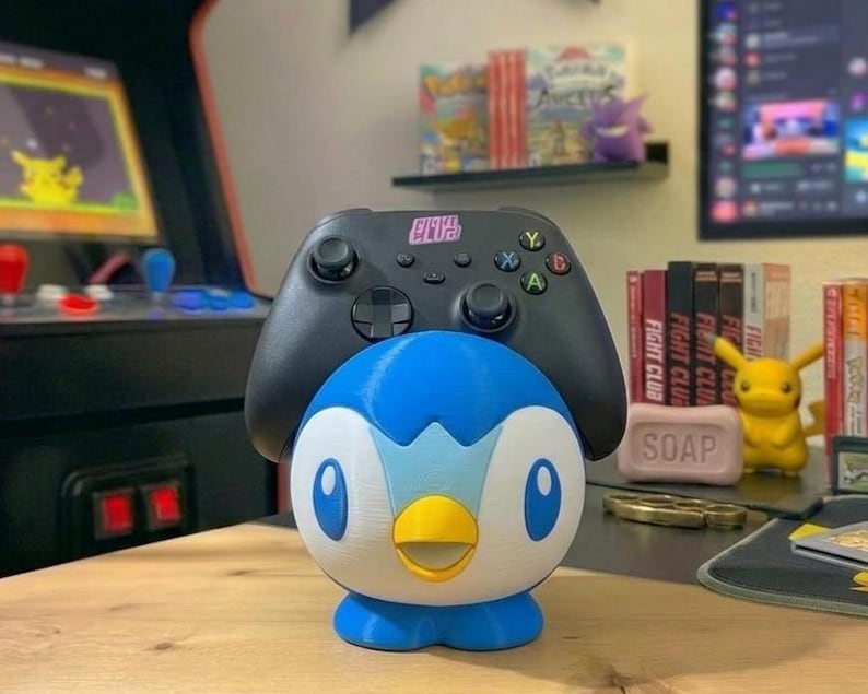 K&ouml;nnte beinhalten: Ein blau-wei&szlig;-gelber Piplup-Controllerhalter mit einem schwarzen Xbox-Controller obenauf. Der Halter steht auf einer Holzoberfl&auml;che, mit einem Retro-Arcade-Spiel und anderem Gaming-Zubeh&ouml;r im Hintergrund.