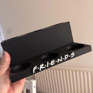 May include: A black wooden shelf with four circular cutouts and the word "FRIENDS" in white letters. The shelf has a rectangular design with a raised back section and is held by a hand.