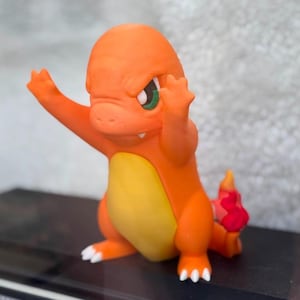May include: A small orange figurine of a bipedal creature with a yellow belly, green eyes, and a small white tooth visible. The creature has its arms raised in a defensive posture and a red flame-shaped detail at the tip of its tail.