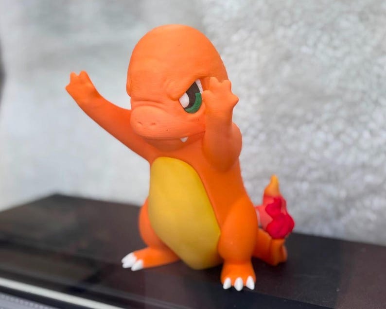 May include: A small orange figurine of a bipedal creature with a yellow belly, green eyes, and a small white tooth visible. The creature has its arms raised in a defensive posture and a red flame-shaped detail at the tip of its tail.