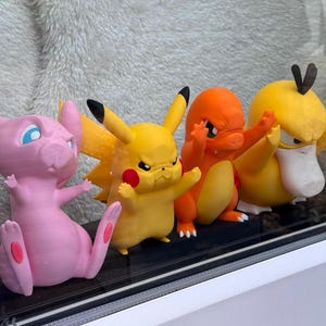 Puede incluir: Un conjunto de cuatro peque&ntilde;as figuras de personajes de pl&aacute;stico alineadas. Las figuras incluyen un Mew rosa, un Pikachu amarillo, un Charmander naranja y un Psyduck amarillo. Cada personaje tiene una expresi&oacute;n facial seria y determinada.