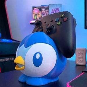 K&ouml;nnte beinhalten: Ein blau-wei&szlig;es Pinguin-Controller-Halter mit gelbem Schnabel. Ein schwarzer Xbox-Controller liegt oben auf. Der Halter steht auf einer schwarzen Oberfl&auml;che, mit einem pinkfarbenen Mauspad und einem Seifenst&uuml;ck.