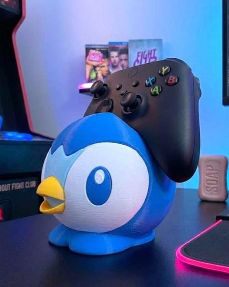K&ouml;nnte beinhalten: Ein blau-wei&szlig;es Pinguin-Controller-Halter mit gelbem Schnabel. Ein schwarzer Xbox-Controller liegt oben auf. Der Halter steht auf einer schwarzen Oberfl&auml;che, mit einem pinkfarbenen Mauspad und einem Seifenst&uuml;ck.
